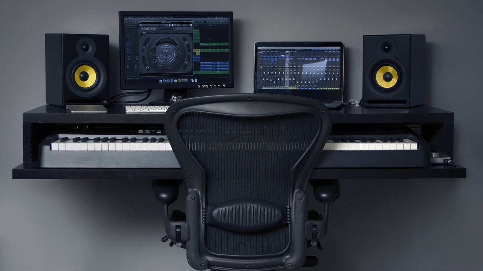 Image shows two computer screens, some speakers and a keyboard on a desk and an office chair in front of it.