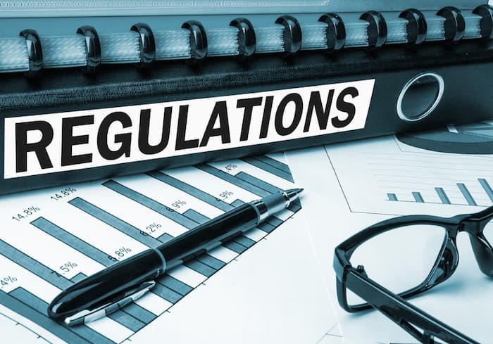 Image shows a close up image of desk, with a folder that reads "Regulations" on it's spine, as well as some sheets of paper, a pen and some glasses.