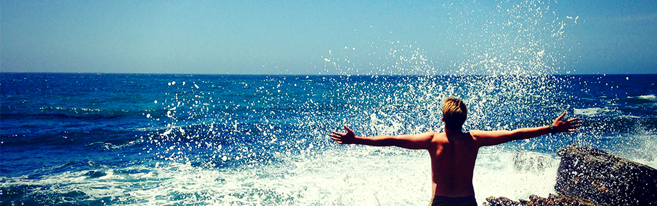 This is an image of someone facing the azure blue sea, arms out and wide shut in a silhouette as a wave crashes over them.