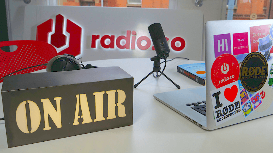 This is the featured image: a lightbox reading ON AIR, a microphone in the middle of the shot, the background being a lit up radio.co logo, and on the right a Macbook emblazoned with stickers.