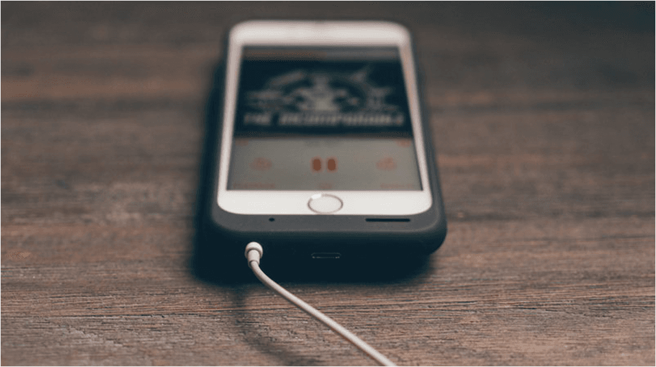 On a dark mahogany backdrop, there is a white iPhone in a case in the middle of the screen. There is indecipherable album art visible, and a wire, presumably headphones, plugged in.