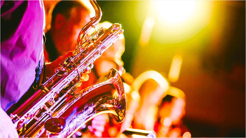 This is a close up video of someone in a pink shirt playing a gold saxophone. In the background are a blurred band. The lights in the venue are yellow.